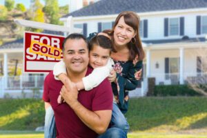 Happy Mixed Race Family In Front of House and Sold For Sale Real Estate Sign.